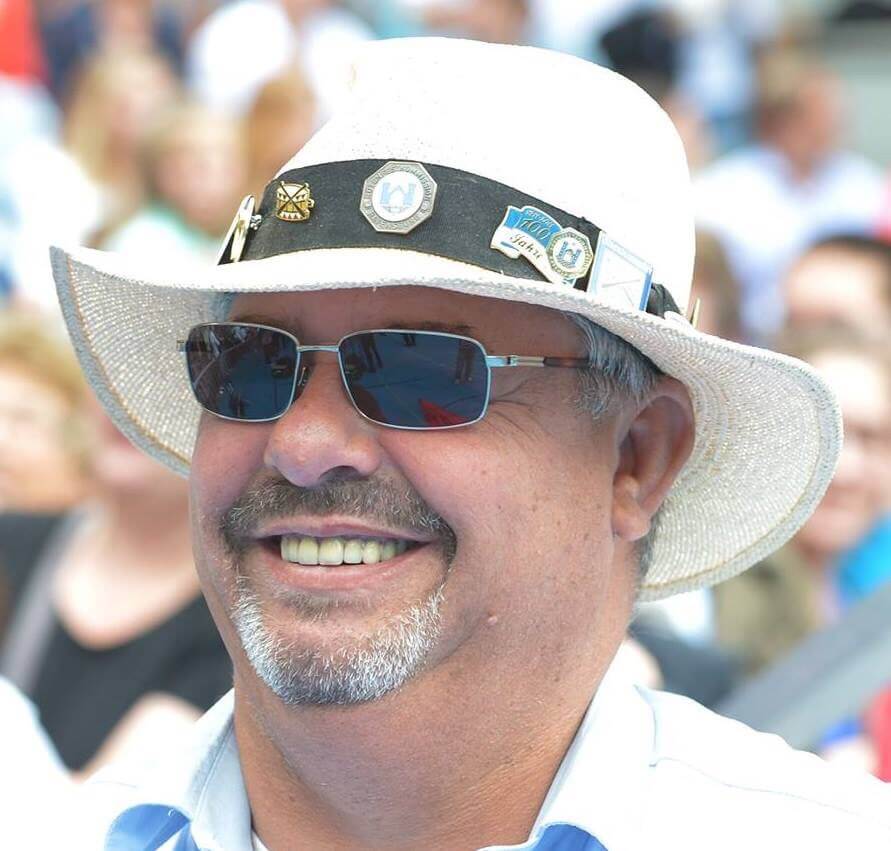 Mann, der einen weißen Hut mit Anstecknadeln, eine Sonnenbrille und ein hellblaues Hemd trägt und im Freien lächelt, mit einer Menschenmenge im verschwommenen Hintergrund.