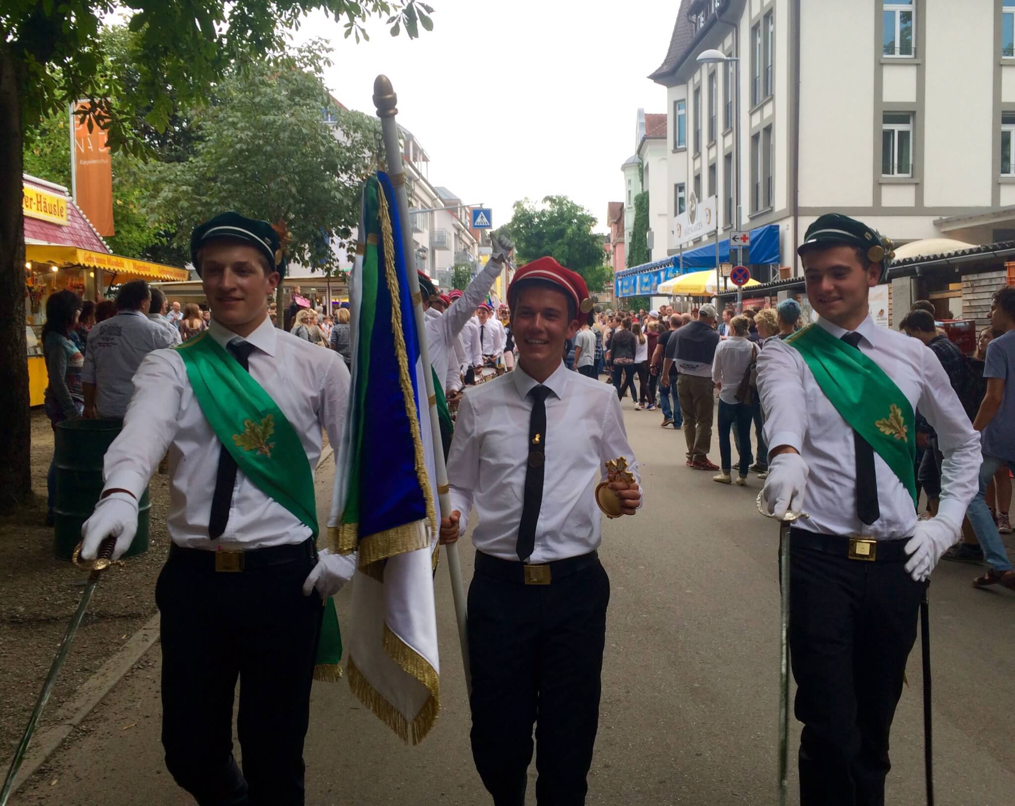 Drei junge Männer in weißen Hemden, schwarzen Hosen und grünen Schärpen marschieren in einer Straßenparade, einer von ihnen mit einer Fahne in der Hand, während Schaulustige am Rande einer Stadtstraße zusehen.