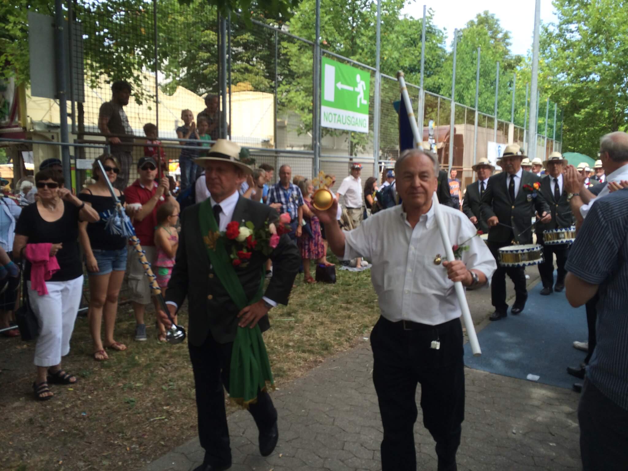 Ein Mann in einem weißen Hemd hält einen gelben Ball und eine weiße Stange und führt an einem sonnigen Tag eine Parade mit Männern in Anzügen und Schaulustigen hinter einem Zaun an.
