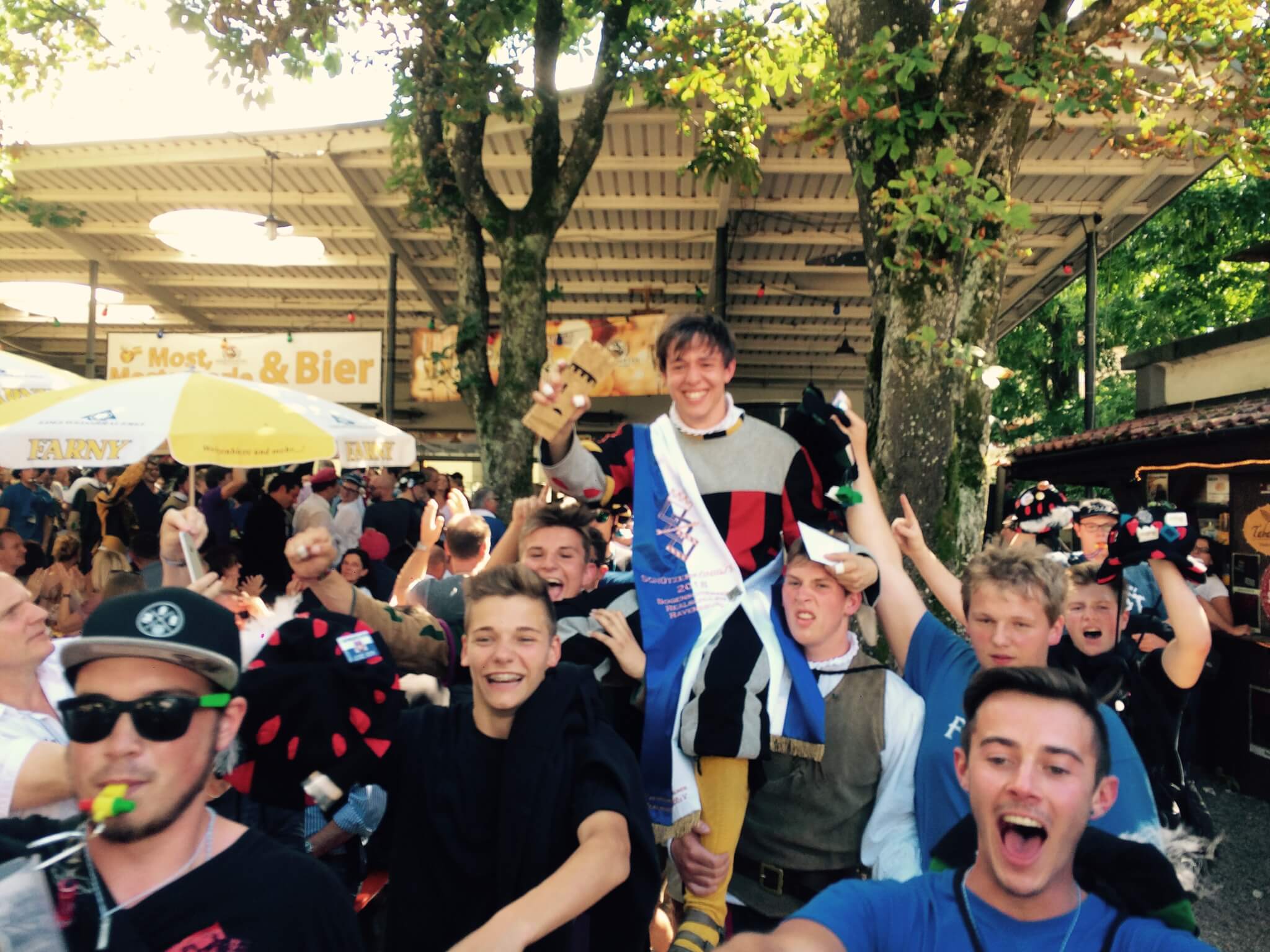 Eine Gruppe junger Männer feiert bei einer Veranstaltung im Freien, einer hält eine Trophäe und trägt eine Schärpe, umgeben von jubelnden Menschen unter einem Baldachin und Bäumen.