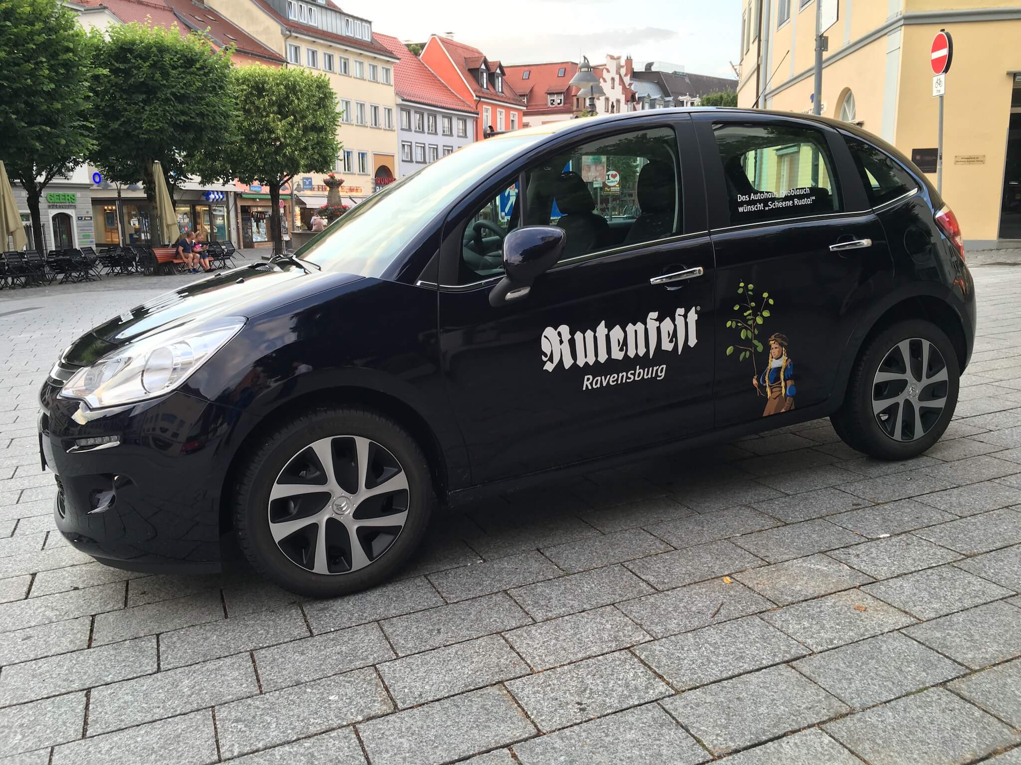 Schwarzer Kleinwagen mit der Aufschrift "Nutenheit Ravensburg" und einer kleinen Grafik an der Seite, geparkt auf einer Kopfsteinpflasterstraße in einer europäischen Stadt.