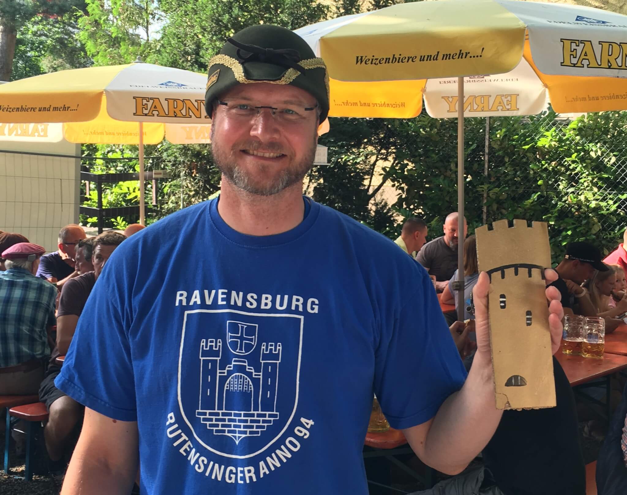 Ein Mann mit einem blauen "Ravensburg"-T-Shirt und einer Tarnkappe hält einen Burgturm aus Pappe und steht im Freien an einem Tisch unter gelben Regenschirmen.