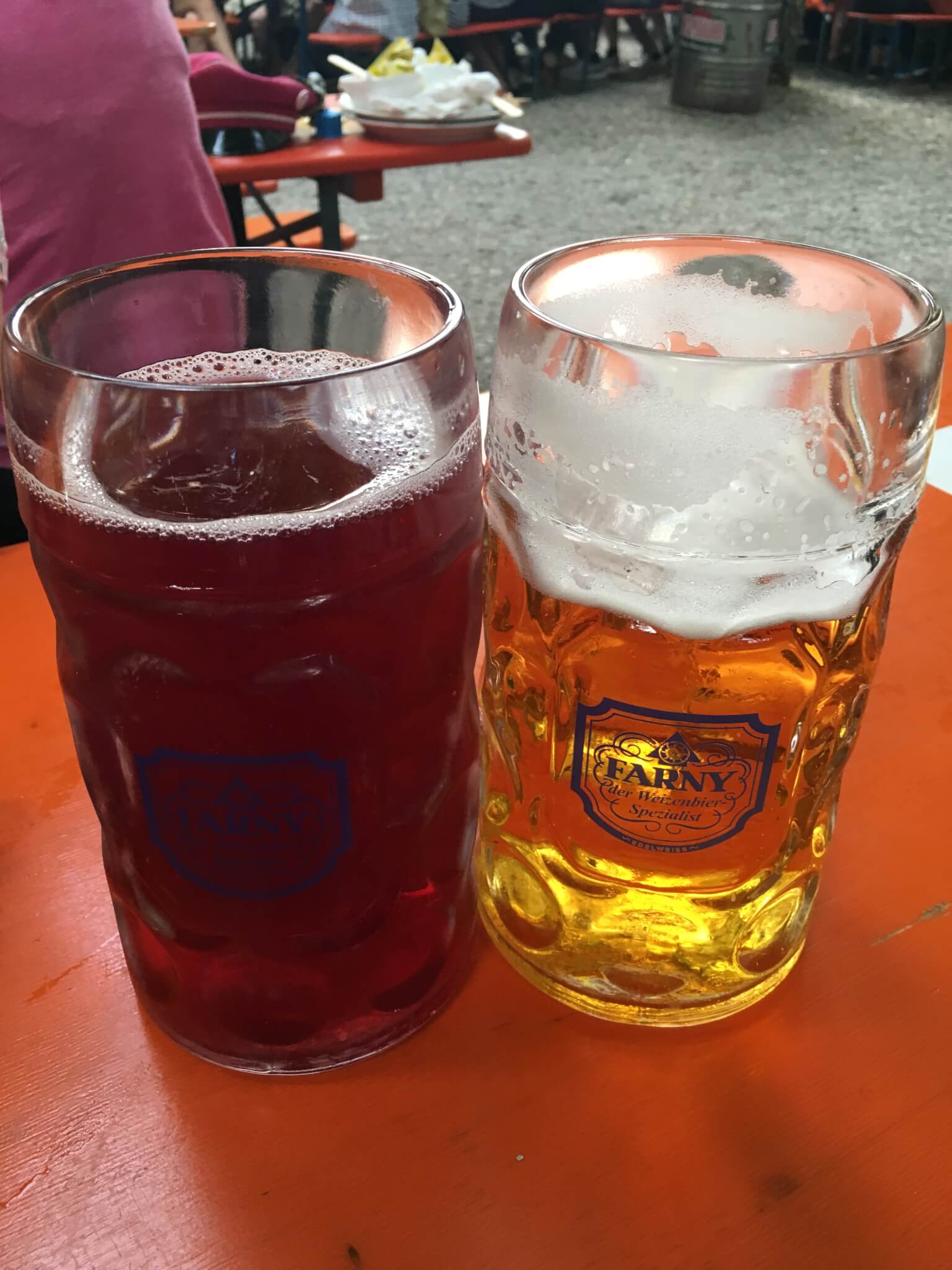 Zwei große Bierkrüge auf einem orangefarbenen Tisch, einer gefüllt mit einem dunkelroten Getränk, der andere mit goldenem Bier, an einem Sitzplatz im Freien.