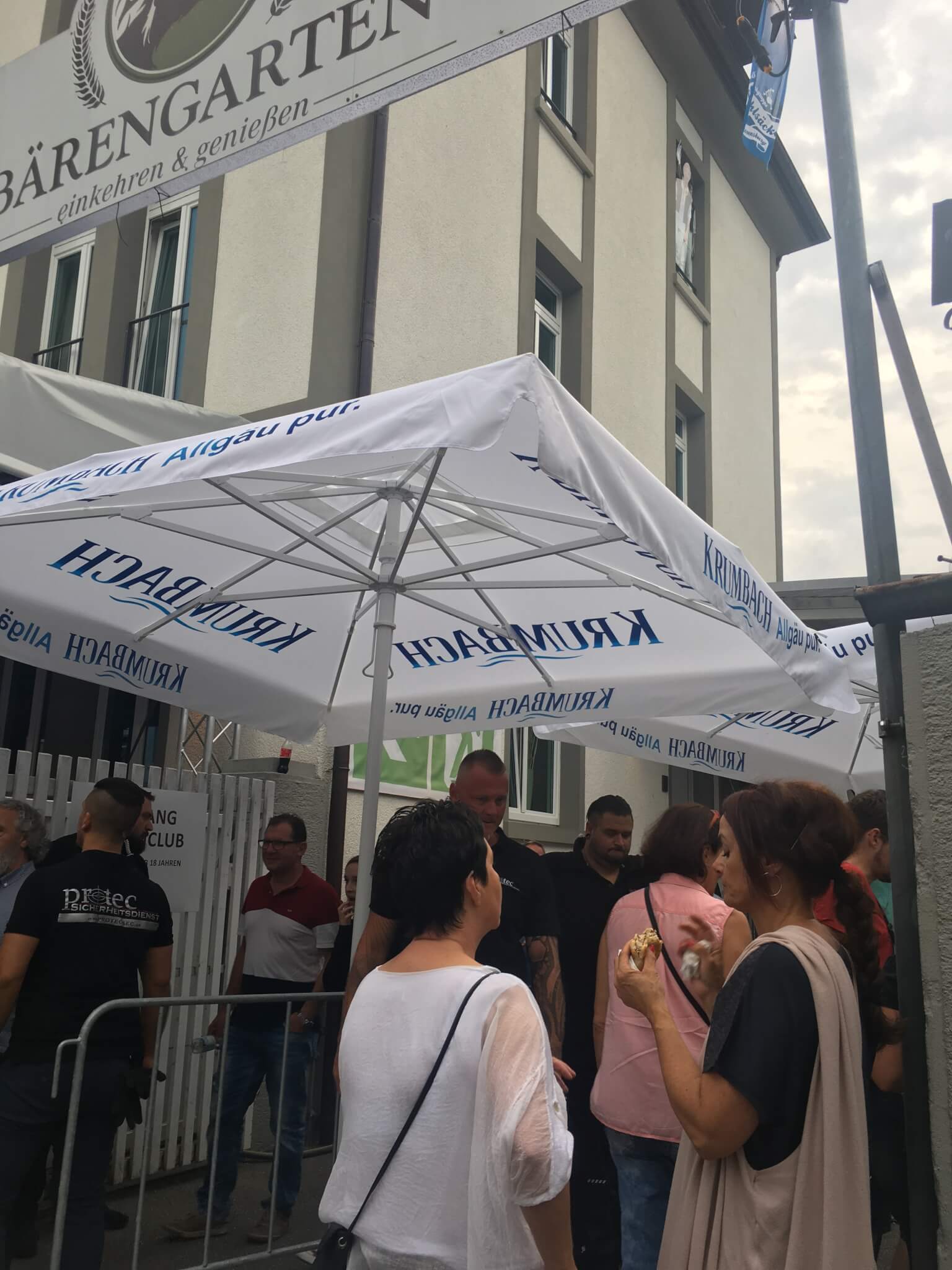 Menschen stehen in einer Schlange vor einem Gebäude unter gebrandeten weißen Regenschirmen in der Nähe eines Schildes mit der Aufschrift "Bärengarten". Der Himmel ist bewölkt und die Gruppe scheint darauf zu warten, hineinzukommen.