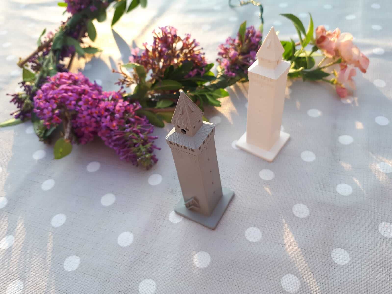 Zwei kleine turmförmige Figuren, eine grau und eine weiß, stehen auf einer gepunkteten Tischdecke neben einem Kranz aus lila und rosa Blumen.