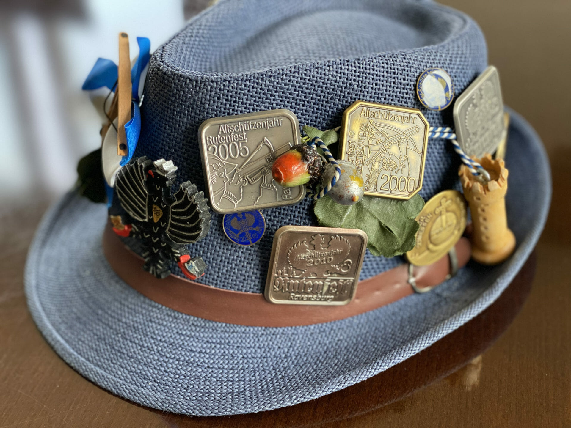 Ein grauer Filzhut, verziert mit verschiedenen metallischen Anstecknadeln, Abzeichen und Ornamenten, darunter Medaillen mit Daten und Emblemen, eine Feder und ein Miniatur-Holzturm.