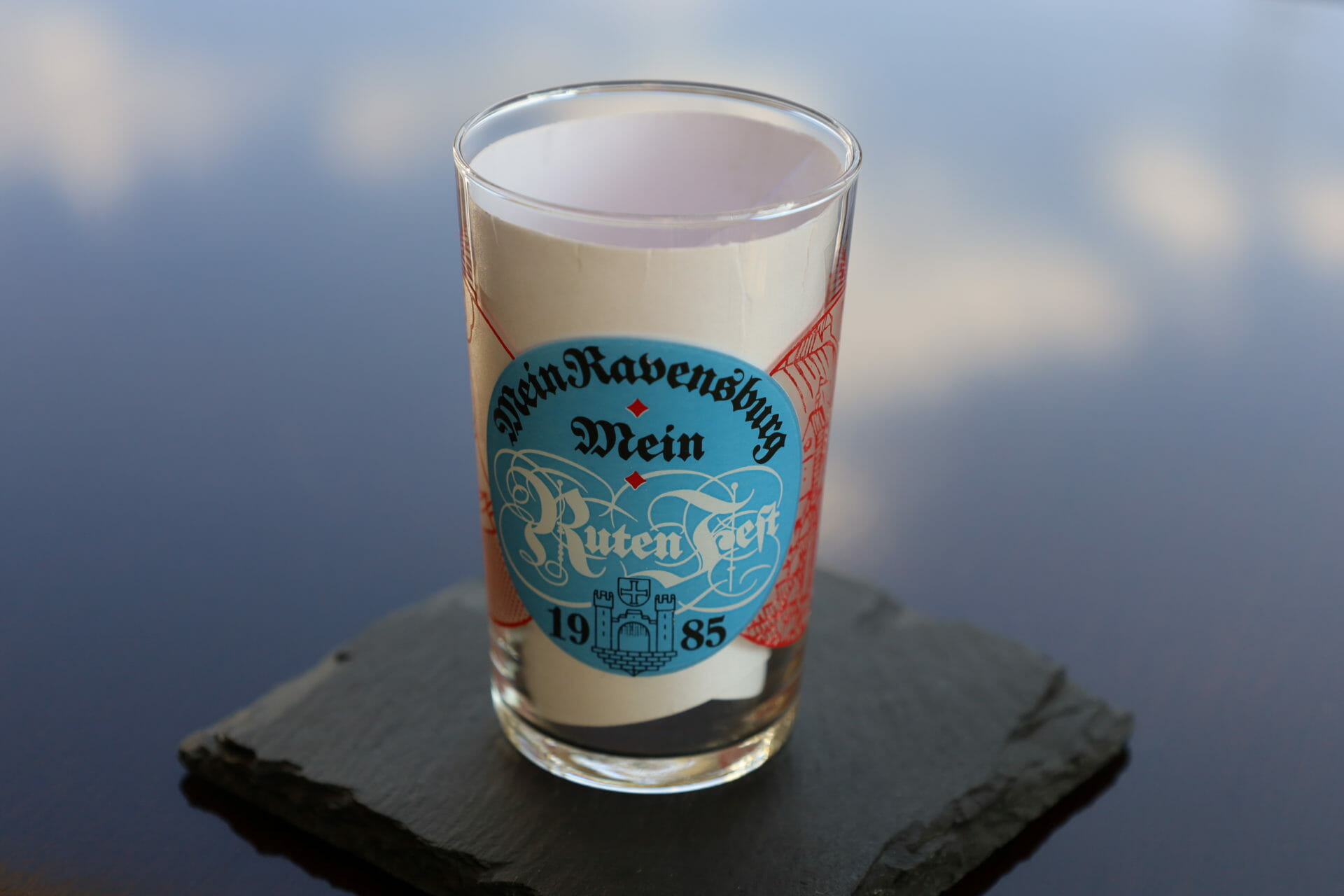 Auf einem dunklen Schieferuntersetzer steht ein Glas mit der Aufschrift "Mein Ravensburger Wein Ruten Fest 1985" mit blauer und roter Schrift und einer Burggrafik.