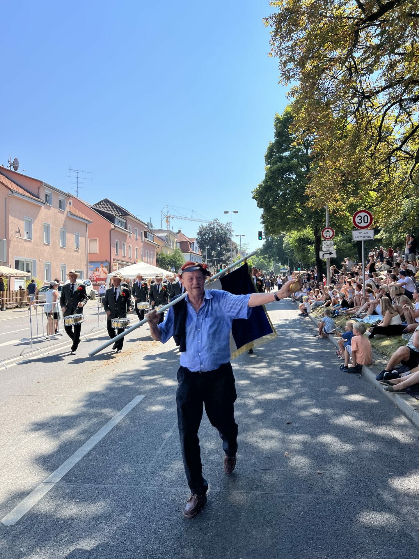 Ein Mann geht mit einer Fahne in der Hand eine Straße entlang, während eine Parade im Freien stattfindet, bei der Musiker marschieren und eine Menschenmenge vom Bürgersteig aus zusieht.