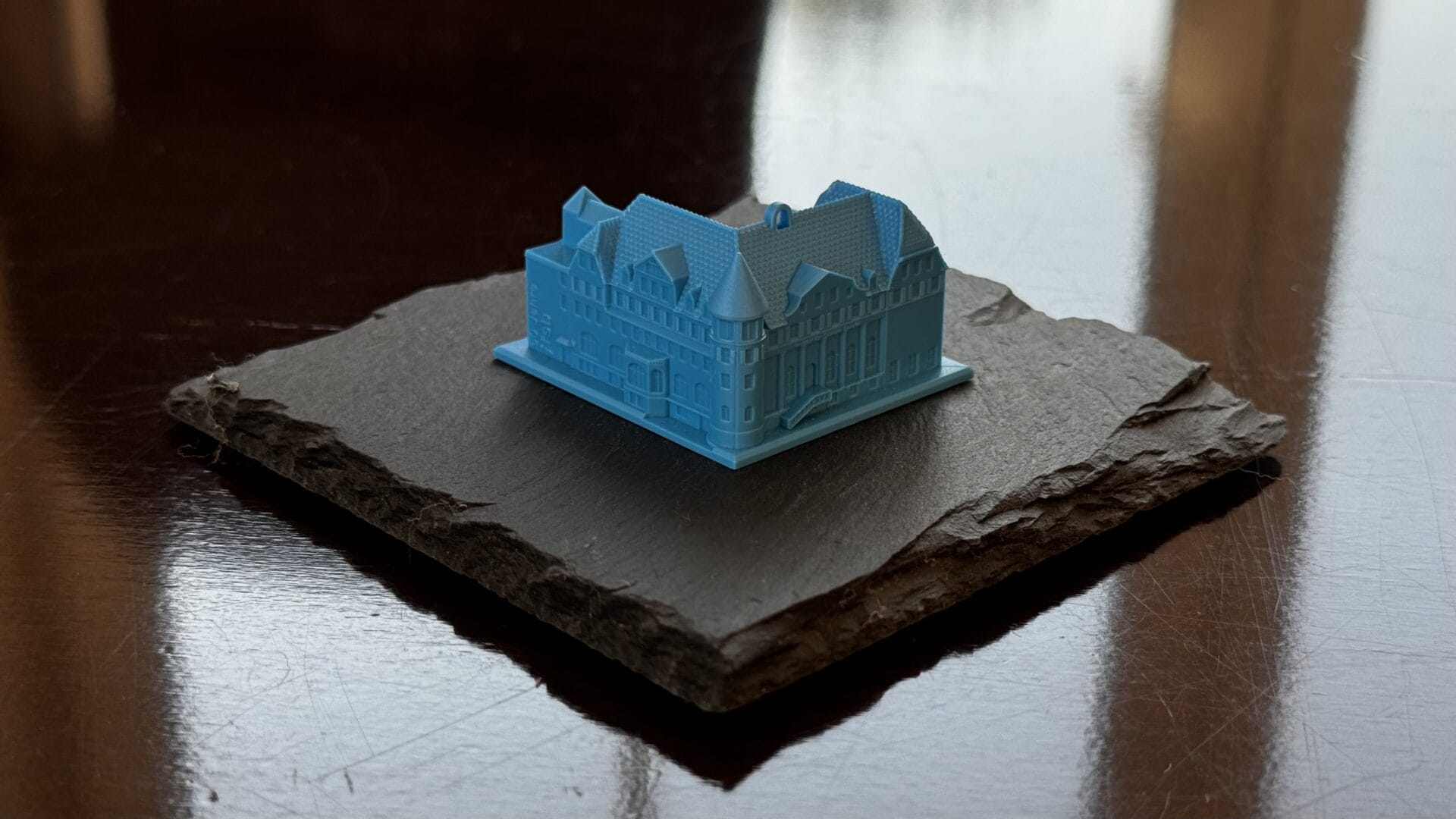 Ein kleines blaues Gebäudemodell sitzt auf einem quadratischen Stück dunklen Steins, das auf einer spiegelnden braunen Fläche steht.