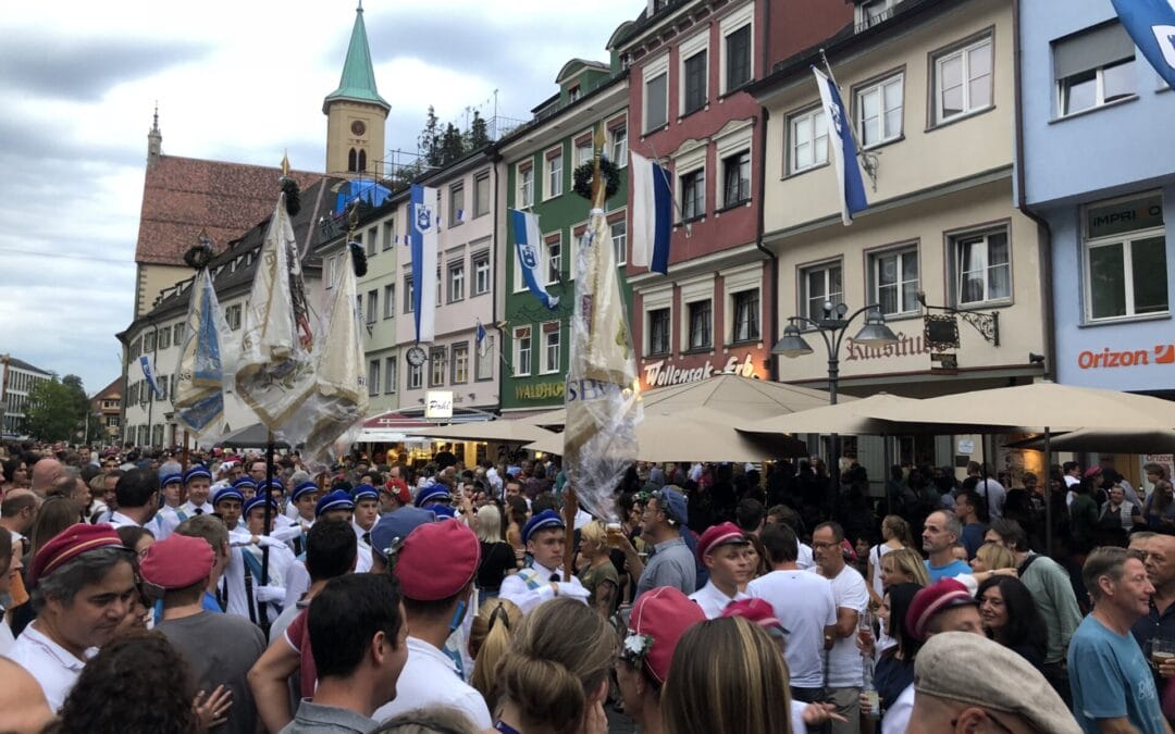Eine große Menschenmenge versammelt sich auf einer von bunten Gebäuden gesäumten Straße, einige Menschen in Uniformen und mit Hüten tragen geschmückte Fahnen; darüber hängen blaue und weiße Flaggen.