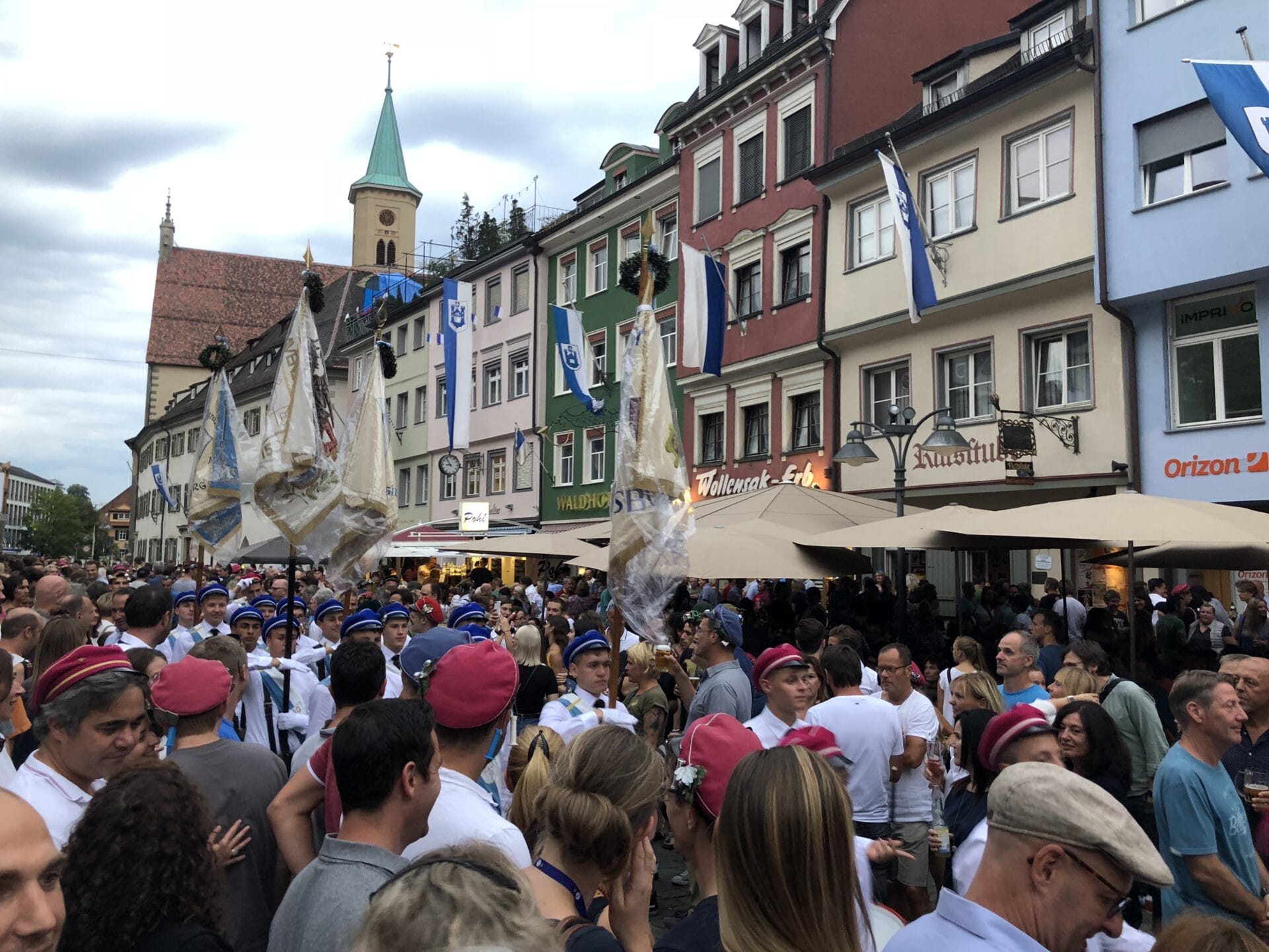 Eine große Menschenmenge versammelt sich auf einer von bunten Gebäuden gesäumten Straße, einige Menschen in Uniformen und mit Hüten tragen geschmückte Fahnen; darüber hängen blaue und weiße Flaggen.